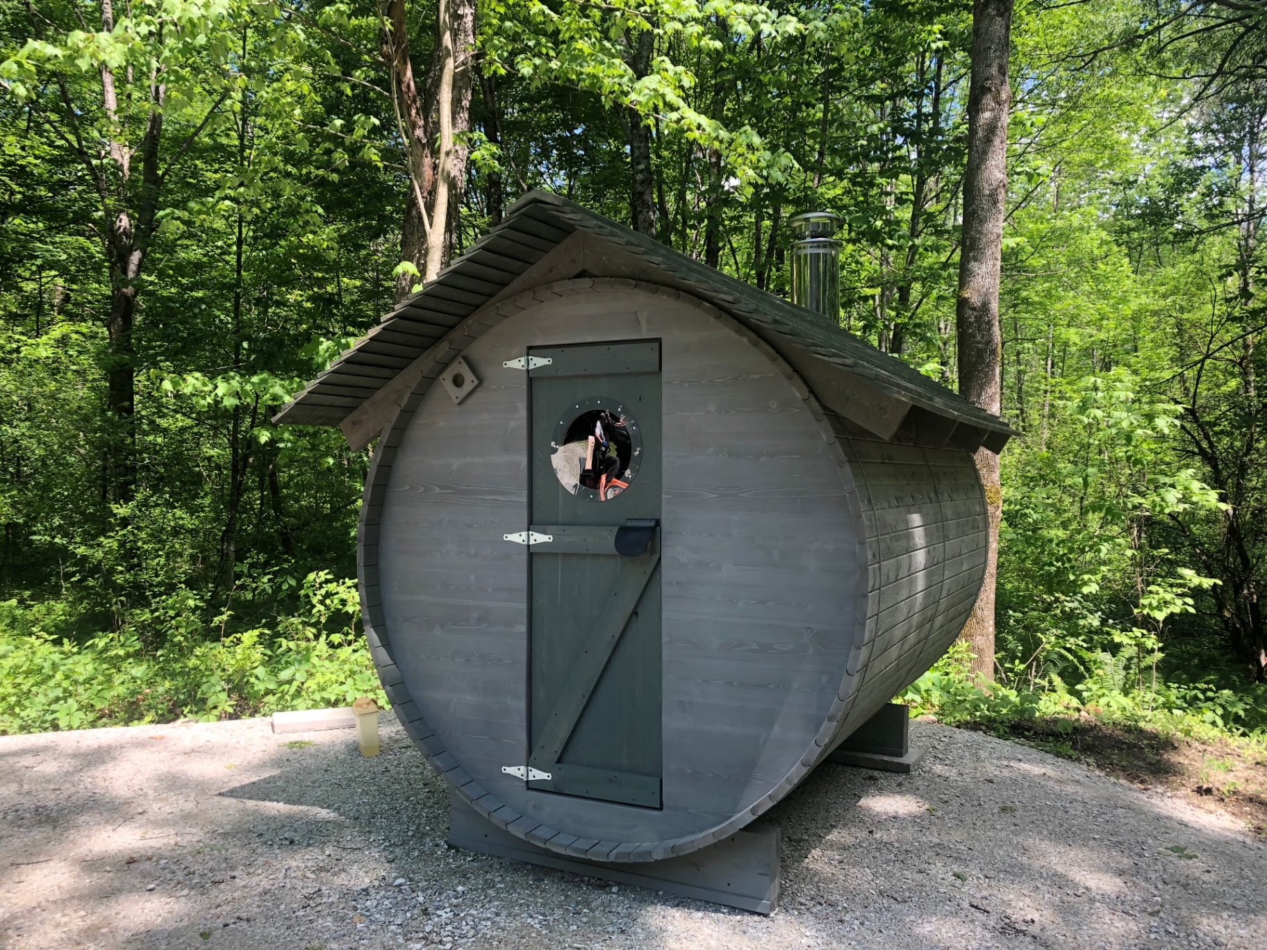 Sauna baril gris installé en forêt avec hublot rond